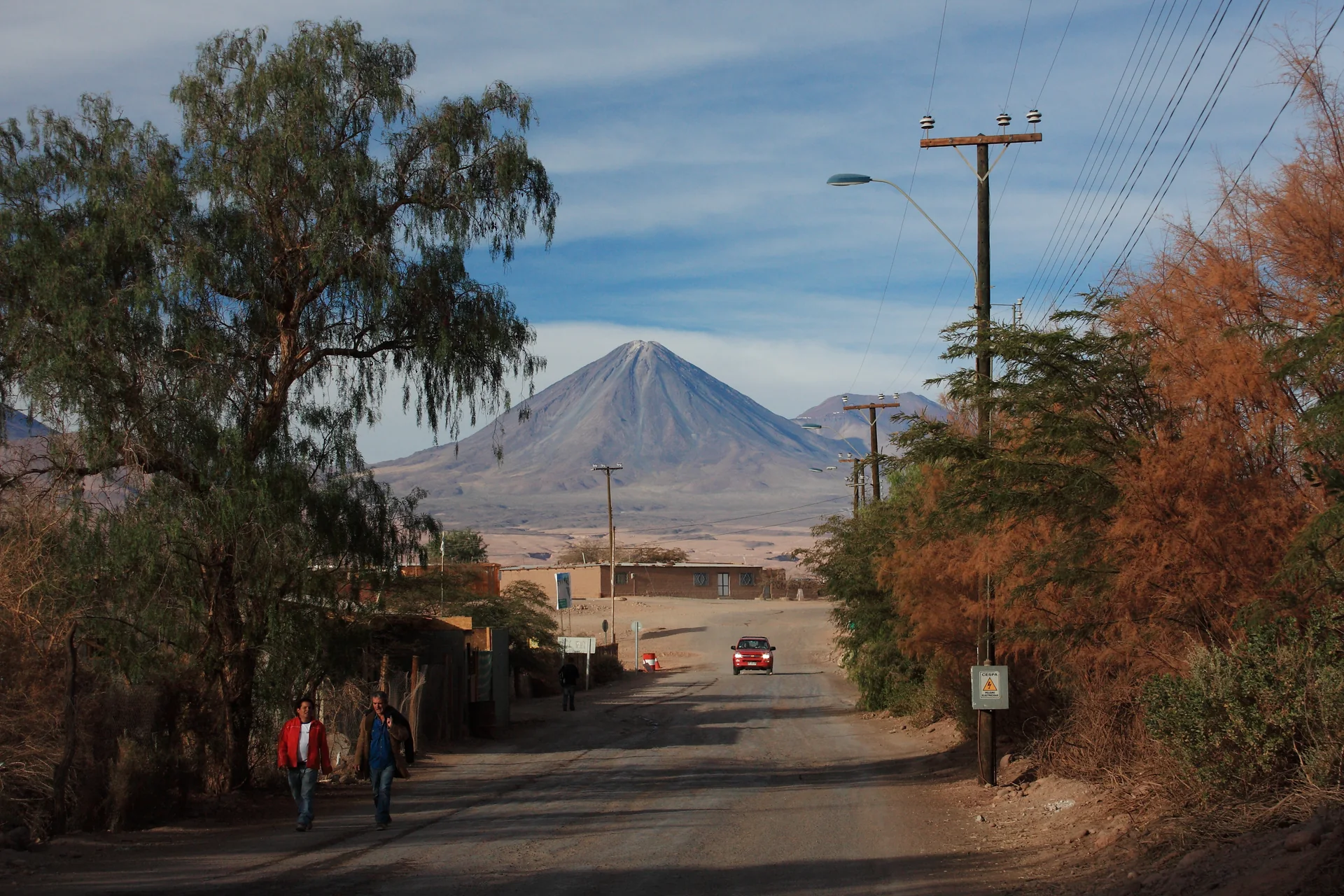 Vista diurna de San Pedro de Atacama com casario baixo, vegetação do oásis, cordilheira ao fundo e céu azul claro.